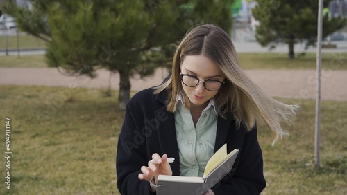 smiling young girl in glasses sitting outdoors in park and making notes in notebook