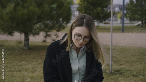 smiling young girl in glasses sitting outdoors in park and making notes in notebook