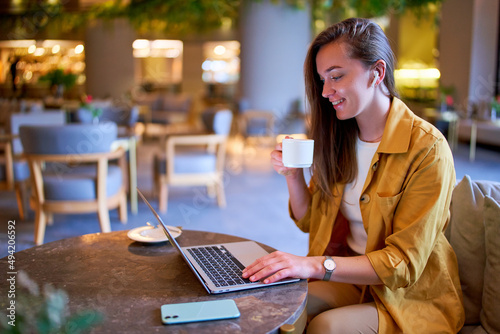Happy cute joyful modern satisfied smart young casual girl freelancer wearing wireless headphones works remotely at laptop in a cafe