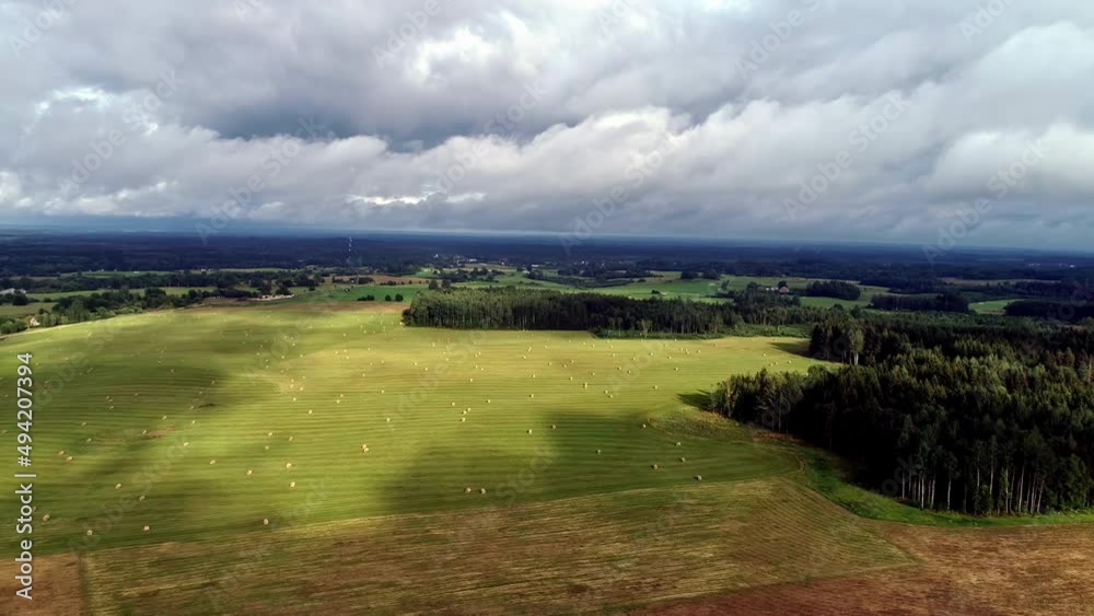 Aerial drone shot flying over a raked field with hay bales scattered out on a cloudy day with sun rays falling on the field at daytime. Aerial drone shot flying over a raked field with hay bales scattered out on a cloudy day with sun rays falling on the field at daytime.