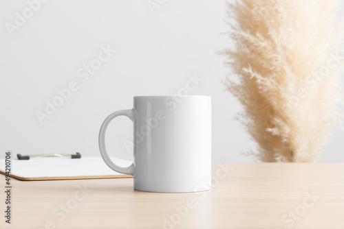 White mug mockup with a clipboard on the wooden table with a pampas decoration.