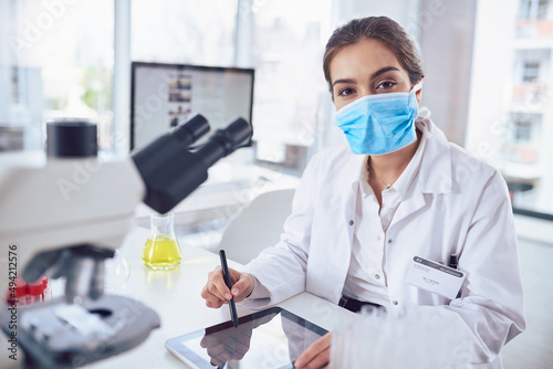 Just another day in the lab. Portrait of a confident young female scientist wearing a surgical mask and working on a digital tablet while looking at the camera in a laboratory.