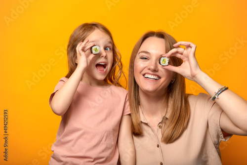 Funny mom with daughter holding sushi rolls in front of eyes on yellow background