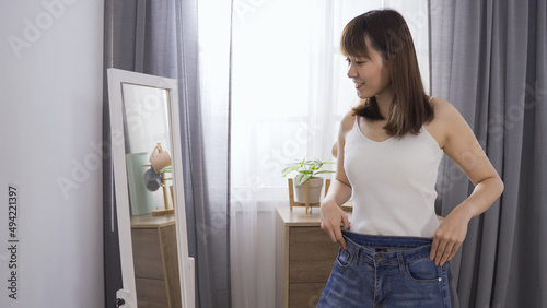 slim asian girl feeling happy about being able to fit into her jeans and standing in front mirror during clothes changing time in the morning.