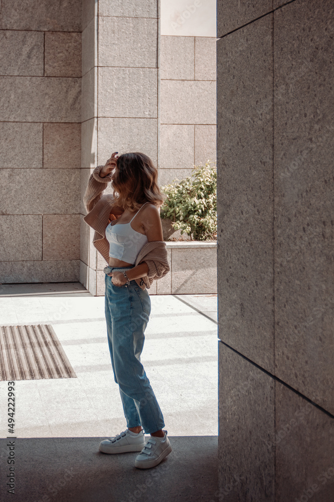 young curly brunette girl in white top and blue jeans is standing in profile near store column at sunny day and looking with flirt. lifestyle concept, free space