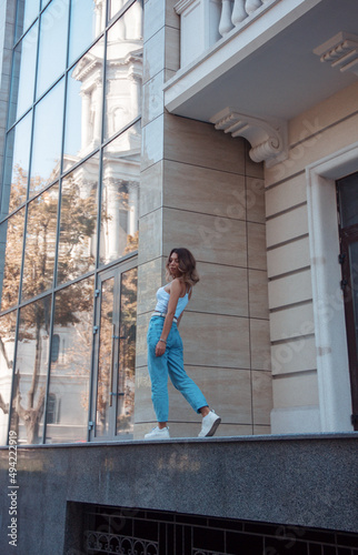 young curly brunette girl in white top and blue jeans is standing casual in step from the back and looking away near modern glass building background at city. lifestyle concept, free space