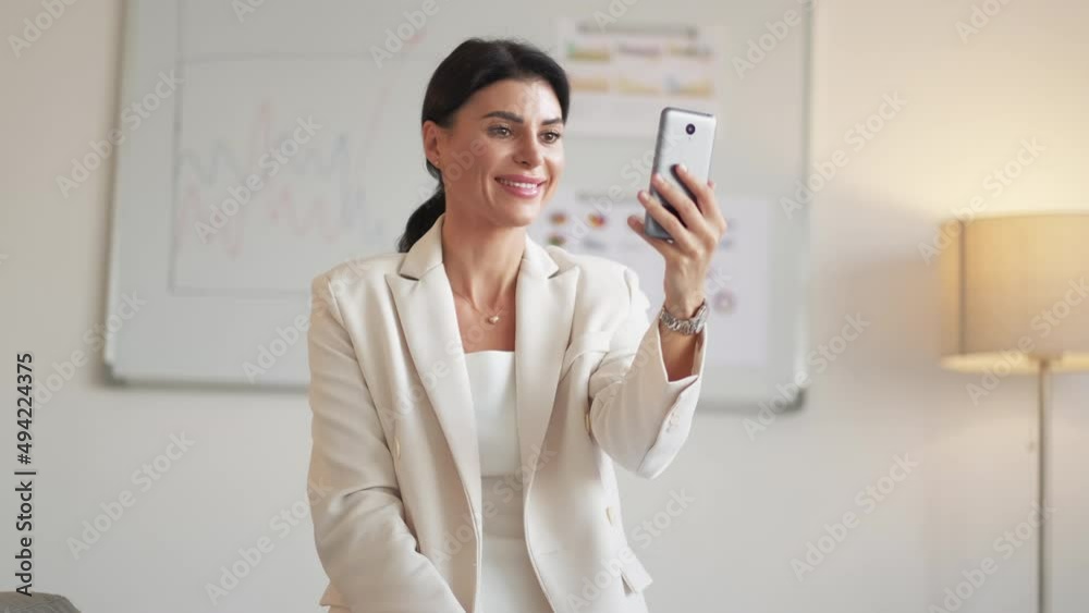 Online meeting. Flirting woman. Successful life. Happy feminine lady in elegant suit talking on camera smartphone on office background in light room interior. Online meeting. Flirting woman. Successful life. Happy feminine lady in elegant suit talking on camera smartphone on office background in light room interior.