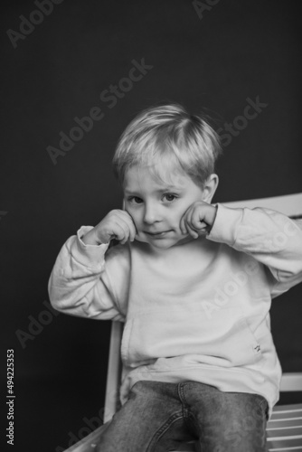 brunette kid in white pullover sitting on white chair against black studio background. Childhood, fashion. Close up, copy space