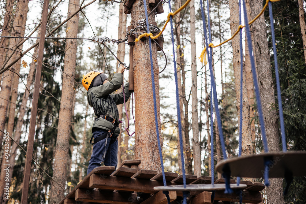 Teenager boy in safety equipment routing and climbing in adventure rope park. Child in helmet climbing trees in an extreme park. Outdoor activities. Active lifestyle