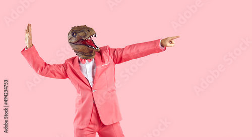 Funny goofy guy having fun in the studio. Happy energetic excited crazy man wearing stylish funky vibrant pink party suit and dinosaur mask dancing and pointing finger away isolated on pink background