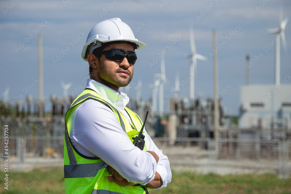 Portrait male engineer wearing white helmet standing in wind turbine power generator station, outdoor background. Portrait male engineer wearing white helmet standing in wind turbine power generator station, outdoor background.