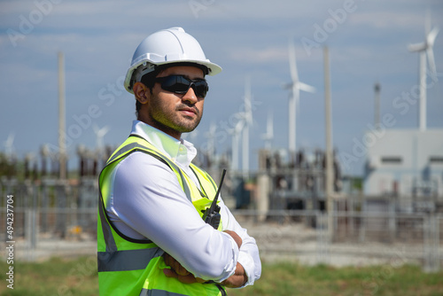 Portrait male engineer wearing white helmet standing in wind turbine power generator station, outdoor background.