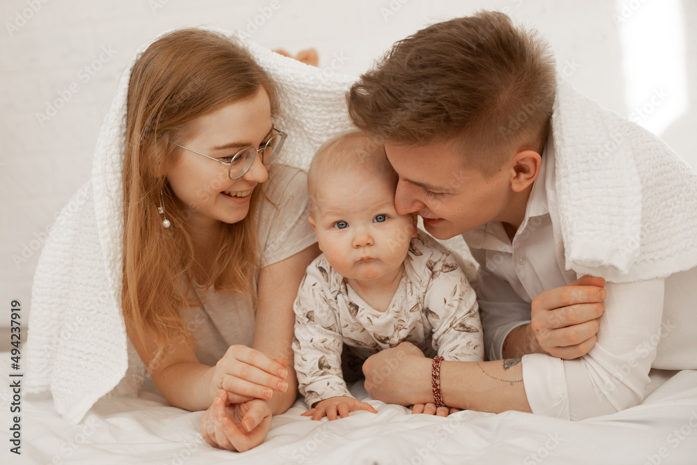 Happy joyful smiling family of woman, man and infant baby lying in white bedspread. Building home of covering towel. Happy joyful smiling family of woman, man and infant baby lying in white bedspread. Building home of covering towel.
