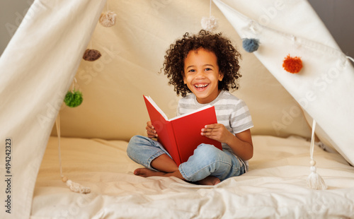 Cute little african american kid curly boy reading book in play tent