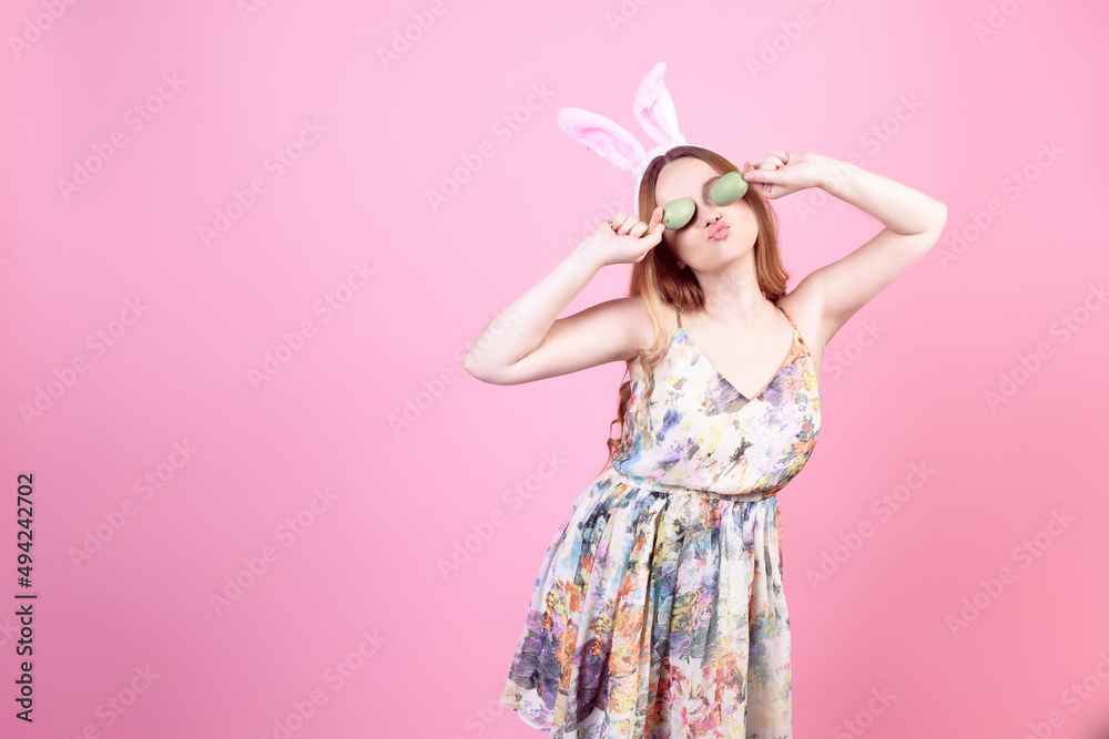Girl in a dress with rabbit ears on a pink background, Easter holidays. Woman beautiful with long hair, easter eggs closes her eyes.
