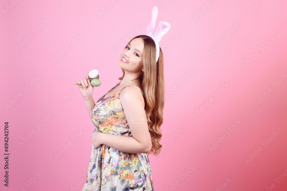 Girl in a dress with rabbit ears on a pink background, Easter holidays. Woman beautiful with long hair, easter eggs, smile and joy