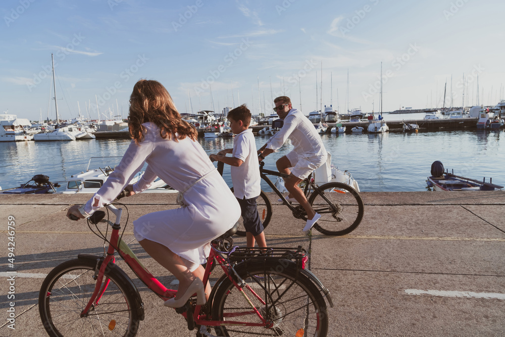 Obraz premium Happy family enjoying a beautiful morning by the sea together, parents riding a bike and their son riding an electric scooter. Selective focus 