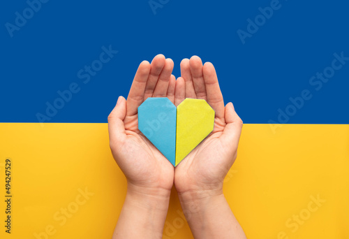 Stand with Ukraine concept. Hands holding blue and yellow heart on Ukraine flag background. Pray for Ukraine peace.