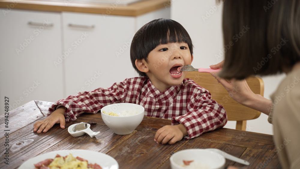 selective focus of cute Asian little boy opening his mouth as his mother feeds him a piece of meat with a fork at dining table. selective focus of cute Asian little boy opening his mouth as his mother feeds him a piece of meat with a fork at dining table.
