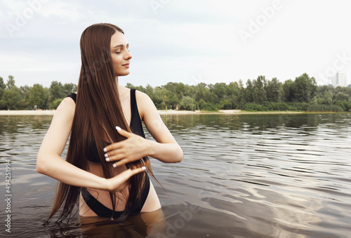 A young woman in a black swimsuit stands in the water in nature in the summer. Swimming and outdoor recreation. Portrait of brunette girl with long hair