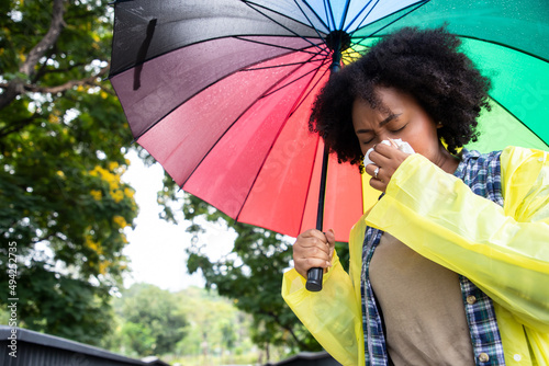 Front view portrait of an African American woman walking towards camera sneezing holding an colorful umbrella under the tree at public park. Health care and insurance concept