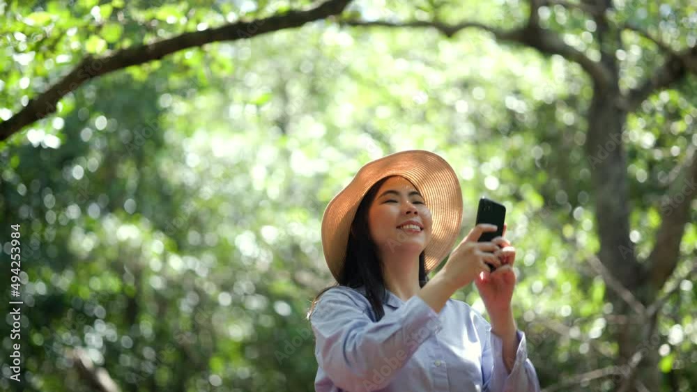 Asian woman travel using cell phone taking video and photo in nature with sunlight. Traveller blogger in mangrove forest. Rest on vacation holiday weekend. Beautiful female with hat and blue shirt Asian woman travel using cell phone taking video and photo in nature with sunlight. Traveller blogger in mangrove forest. Rest on vacation holiday weekend. Beautiful female with hat and blue shirt