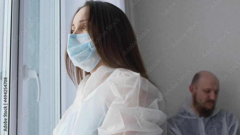 Tired doctors in the rest room for medical staff. A female nurse stands at the window in a surgical gown and a mask, a tired doctor sits behind her, takes off the mask after the operation. Tired doctors in the rest room for medical staff. A female nurse stands at the window in a surgical gown and a mask, a tired doctor sits behind her, takes off the mask after the operation.