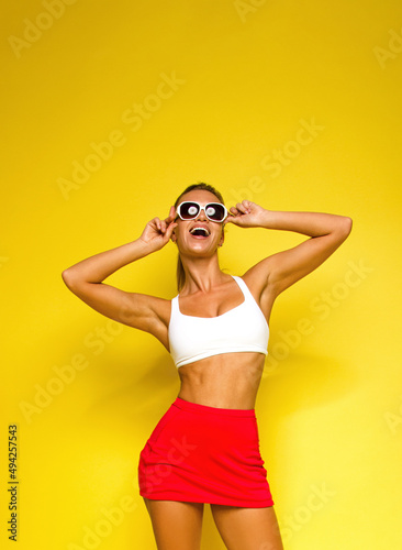 young blonde fit girl in white top and red tennis skirt is standing casual on the yellow wall background and laughing happy with hands near sunglasses. sport lifestyle concept, free space