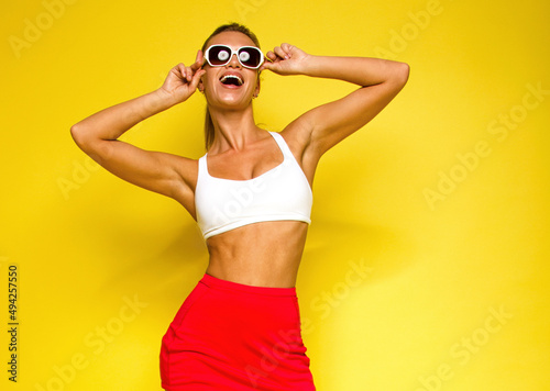 vacation mood. young teen tanned girl is standing on the yellow background and laughing happy with hands near sunglasses. sport lifestyle concept, free space