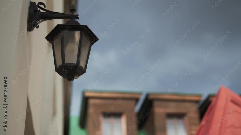 Old black retro lantern with light bulb on the wall of building on the street on cloudy day against the backdrop of house with wooden attic. City decoration in vintage style. Old black retro lantern with light bulb on the wall of building on the street on cloudy day against the backdrop of house with wooden attic. City decoration in vintage style.
