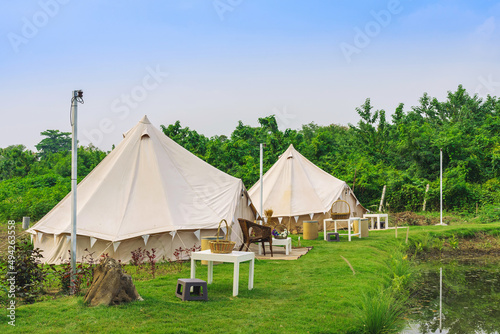 Holiday tents and lounge areas on green lawn place among trees at natural parkland. Camping tent on nature in summer.Travel background. Place for picnic outdoors. Recreation area and camp with tent. © JinnaritT
