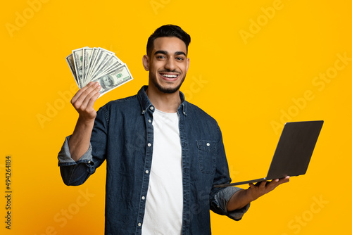 фотографія Positive handsome middle eastern guy showing cash, using notebook