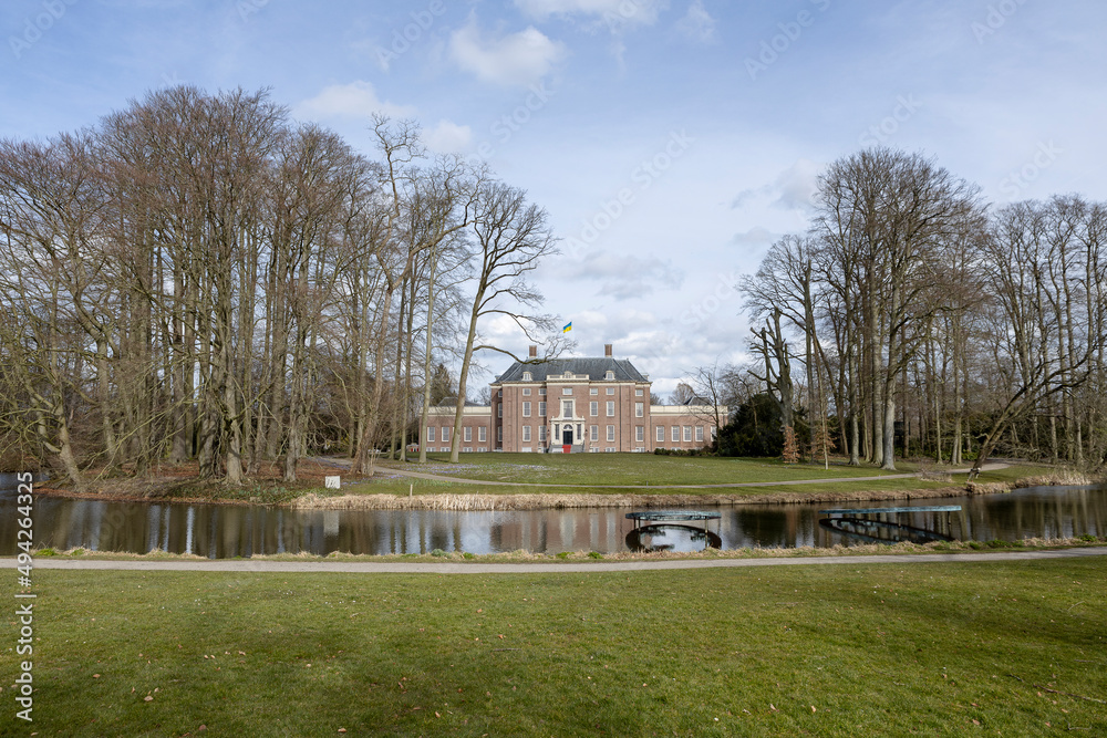 Winter barren trees on a sunny day with park and moat in the foreground and exterior facade of Slot Zeist castle  in the background against a blue sky