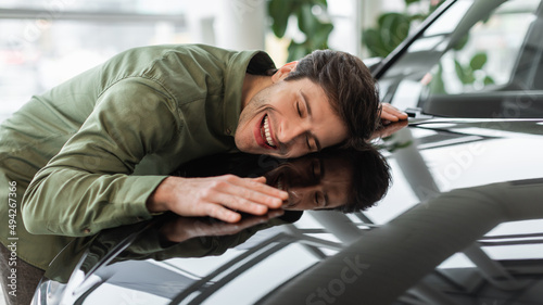 Happy young man hugging hood of his new car, dreaming about buying vehicle at auto showroom, panorama