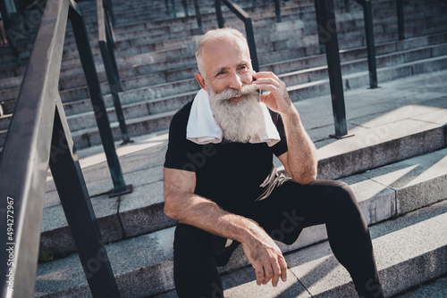 Portrait of attractive bearded cheerful retired grey-haired man working out talking on phone chatting sitting on stone stairs outdoors © deagreez