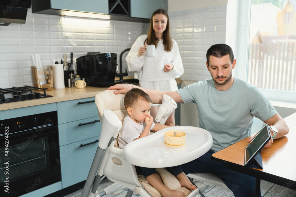 Young parents father and mother feeding the baby. Child on a highchair in the kitchen. Dad, mom and baby - happy full family Young parents father and mother feeding the baby. Child on a highchair in the kitchen. Dad, mom and baby - happy full family