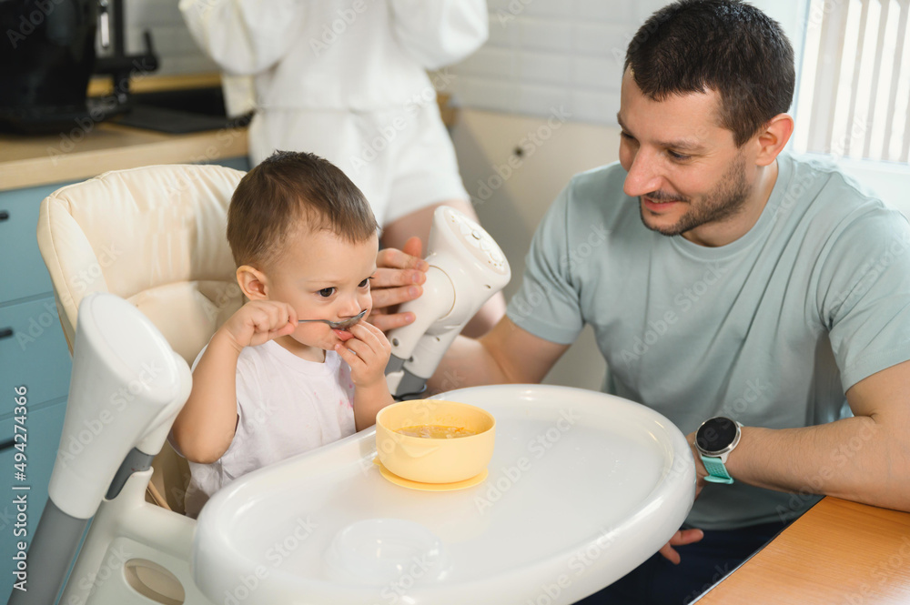 Young parents father and mother feeding the baby. Child on a highchair in the kitchen. Dad, mom and baby - happy full family Young parents father and mother feeding the baby. Child on a highchair in the kitchen. Dad, mom and baby - happy full family