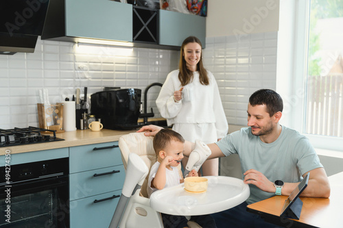 Young parents father and mother feeding the baby. Child on a highchair in the kitchen. Dad  mom and baby - happy full family