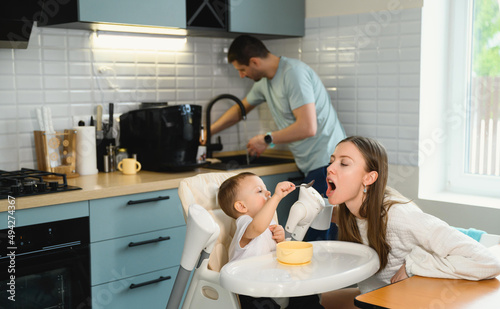 Young parents father and mother feeding the baby. Child on a highchair in the kitchen. Dad, mom and baby - happy full family