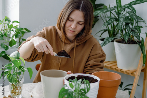 A young caucasian woman gardener holding a shovel and filling a flowerpot with soil preparing it for potting a houseplant sitting surrounded by planted room plants. Indoor or home gardening. 