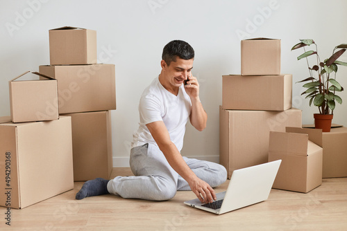 Indoor shot of man wearing white t shirt sitting on floor surrounded with cardboard boxes with belongings and working on laptop and talking phone, works in delivery service online.