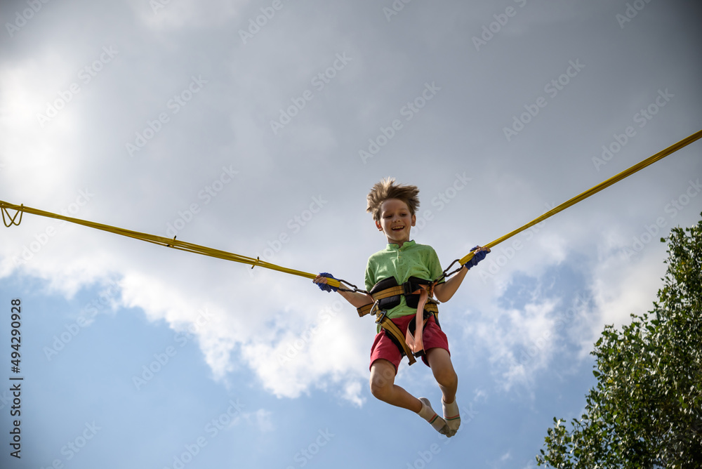 The boy is jumping on a bungee trampoline. A child with insurance and  stretchable rubber bands hangs against the sky. The concept of happy  childhood and games in the amusement park foto