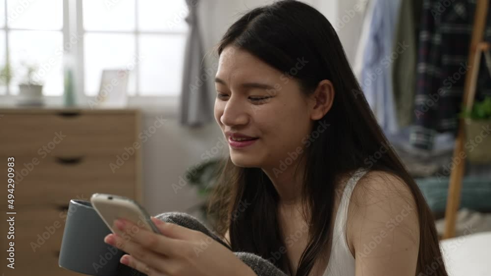 closeup of cheerful Asian young woman is smiling while having fun scrolling on social media on the smartphone and drinking tea in the morning on bed at home closeup of cheerful Asian young woman is smiling while having fun scrolling on social media on the smartphone and drinking tea in the morning on bed at home