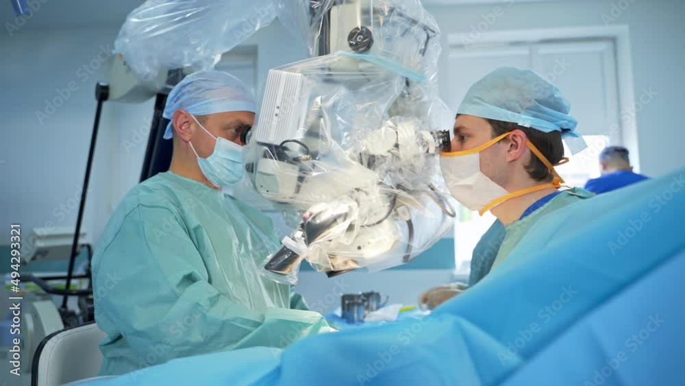 Couple of surgeons stand on both sides of modern equipment looking into binoculars. Hands of doctors with metal instruments move under the apparatus dealing with patient's body. Couple of surgeons stand on both sides of modern equipment looking into binoculars. Hands of doctors with metal instruments move under the apparatus dealing with patient's body.