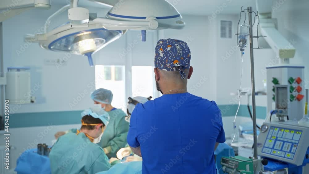Anesthesiologist stands with his back to the camera watching the operation. Team surgeons operating the patient in blur at backdrop. Anesthesiologist stands with his back to the camera watching the operation. Team surgeons operating the patient in blur at backdrop.