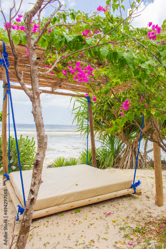Fototapeta premium Hammock on the beach on low tide. Sun bed in tourist resort. Tropical resort. Summer relaxation. Vacations in Africa. Swing with purple flowers on seaside. Exotic nature. Idyllic tropical leisure.