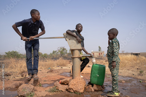Three African children filling up water in a plastic tank at a borehole pump in a rural community in Sub-Saharan Africa. Child labor concept.