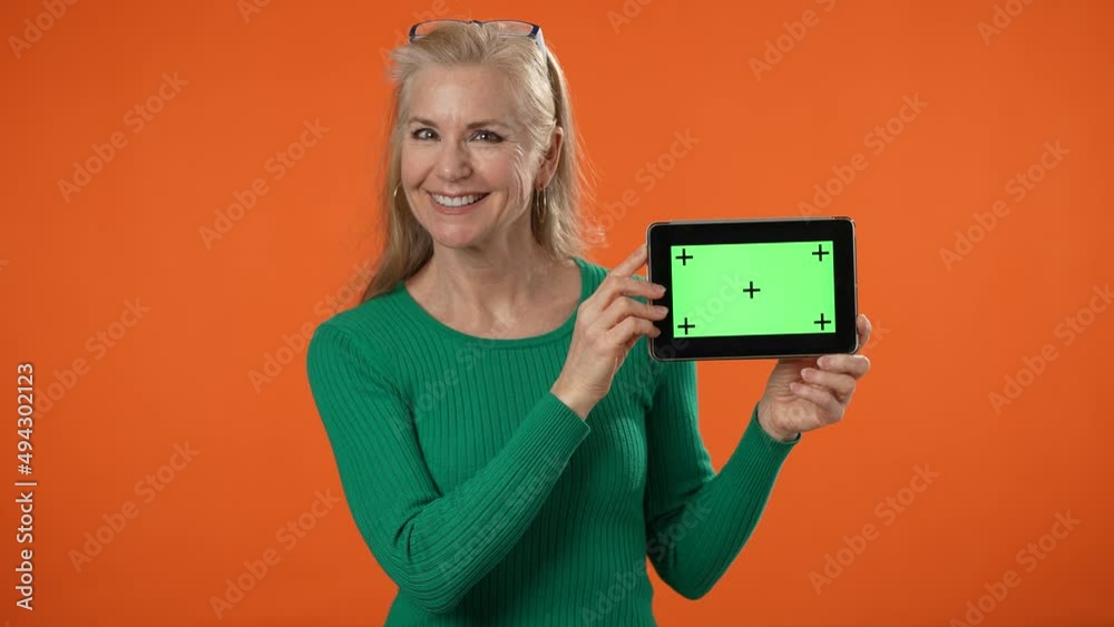 Portrait of smiling mature woman posing with tablet computer with tracking points blank empty green screen, isolated on orange background. People lifestyle concept. Mock up copy space. Portrait of smiling mature woman posing with tablet computer with tracking points blank empty green screen, isolated on orange background. People lifestyle concept. Mock up copy space.