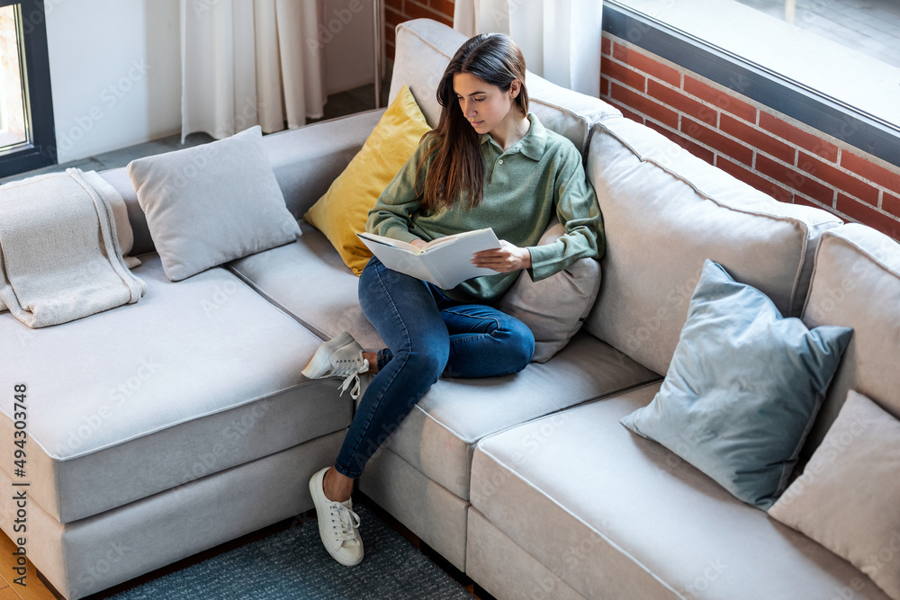 Obraz premium Pretty young woman reading a book while sitting on sofa at home.