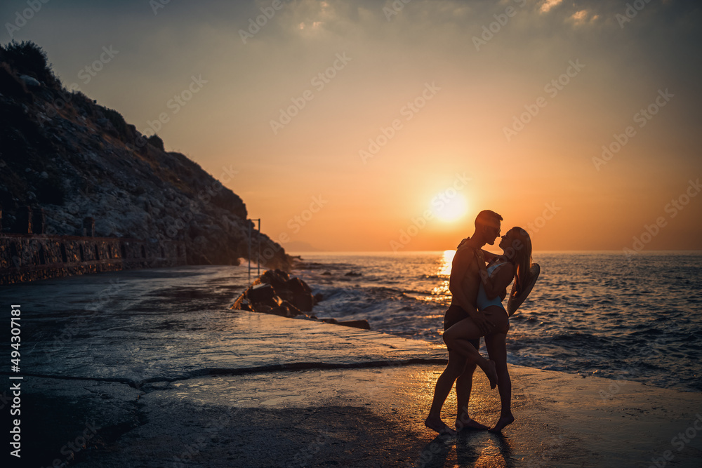 Fototapeta premium Beautiful couple in love on the background of the sunset by the sea. Young woman and man hugging by the sea at sunset
